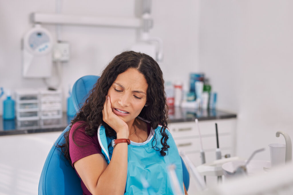 Young mixed race woman sitting in dental chair holding jaw in discomfort. Patient showing signs of tooth pain during consultation,  oral infection or caria. Woman wearing dental bib in clinic with hands on face pressing her cheek, showing discomfort from a toothache.