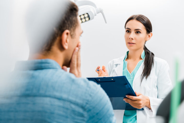 selective focus of female dentist standing with clipboard and talking with african american patient having toothache