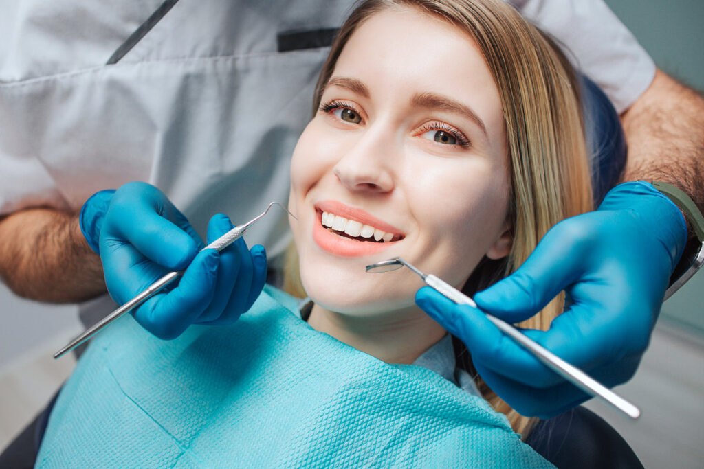 Picture of happy young woman sit in chair and look on camera. She smile and show beautiful white teeth. Doctor hold teeth tools for treatment close to mouth