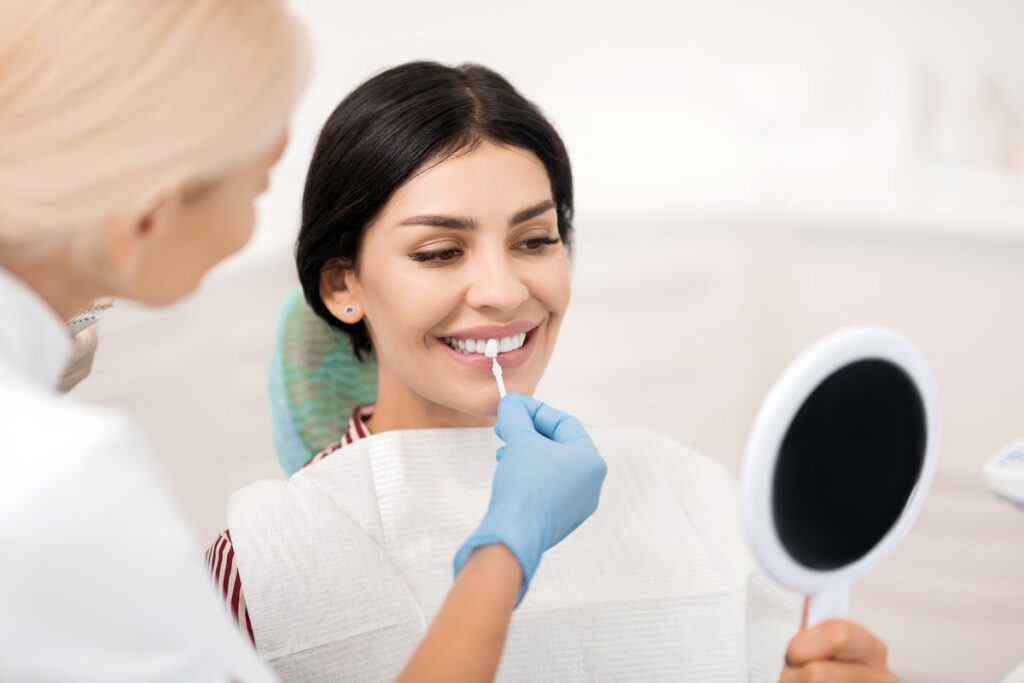 Looking closely. Smilibg woman trying on teeth whitening sample looking in the small mirror at dentists.