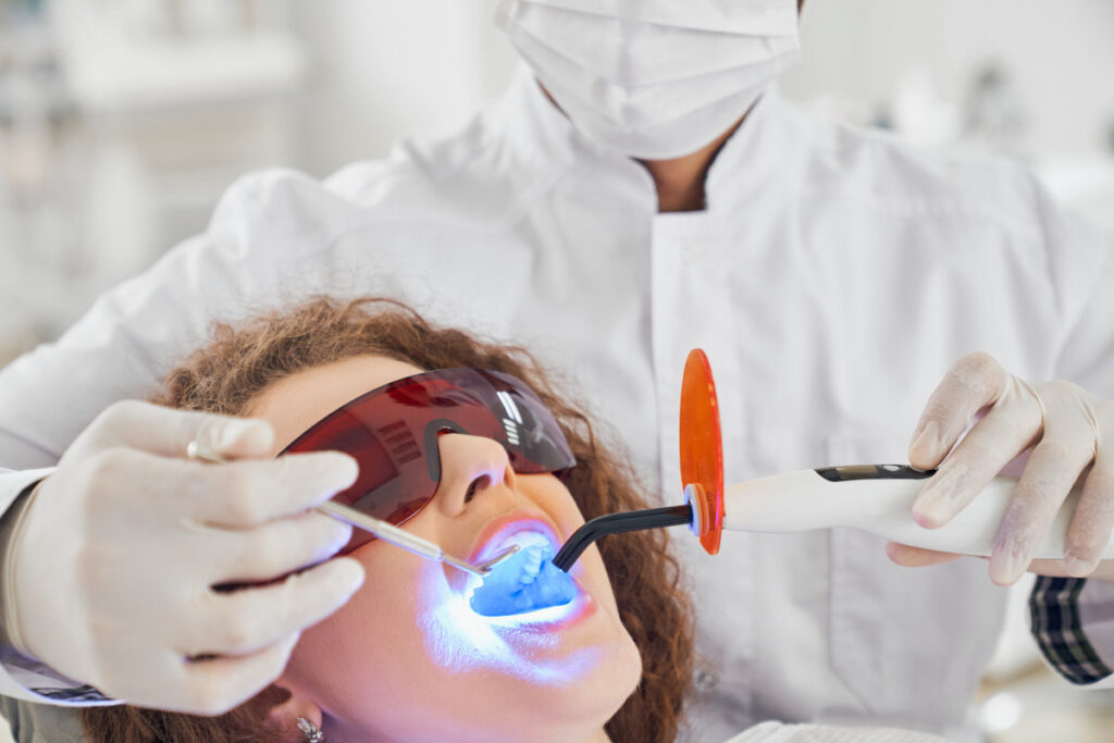 Attractive lady with curly hair lying in dental chair in protective glasses with opened mouth while while male doctor in white glasses doing whitening procedure. Concept of dental care