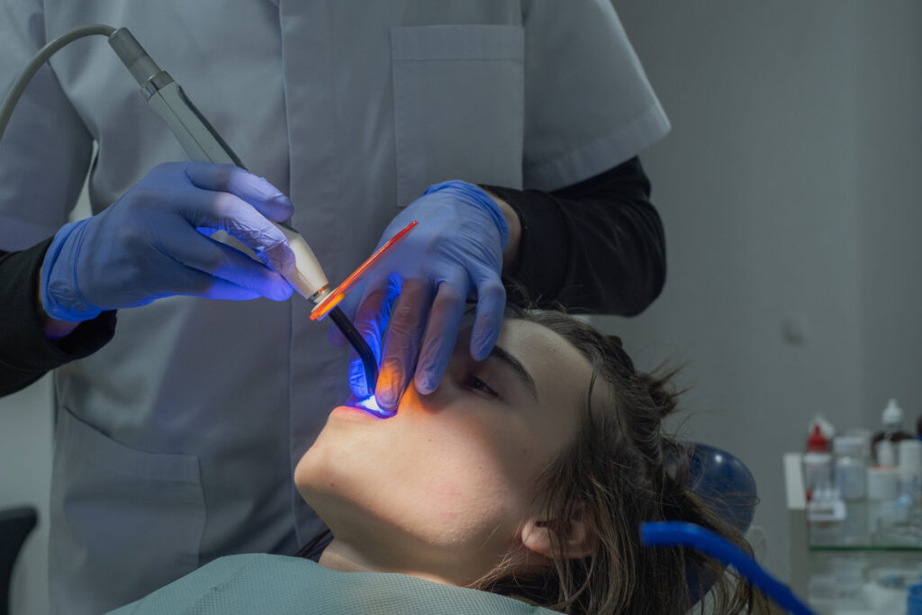 A dentist with blue gloves is using a laser tool to perform a dental procedure on a patient lying in a dental chair. The clinic is well-lit and equipped with dental tools.
