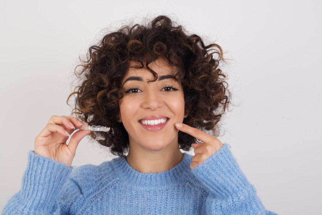 young beautiful caucasian woman wearing blue knitted sweater standing against white studio background holding an invisible aligner braces and smiling. Dental healthcare concept.